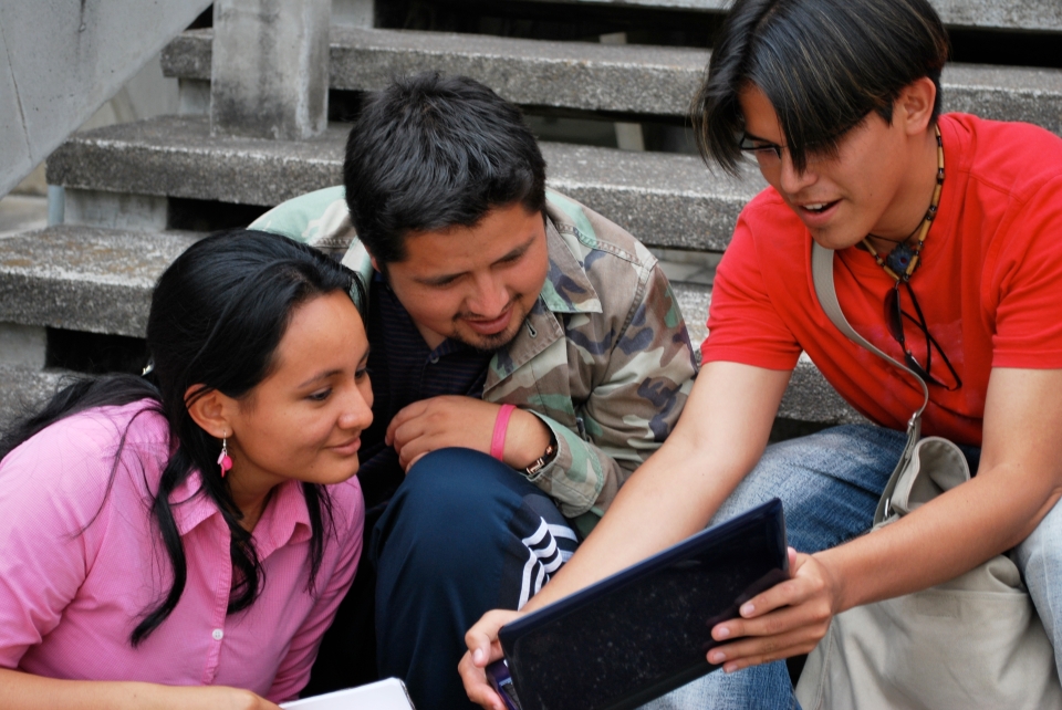 Three Hispanic people sitting on concrete steps, one sharing an image on a laptop which the other two are looking at.