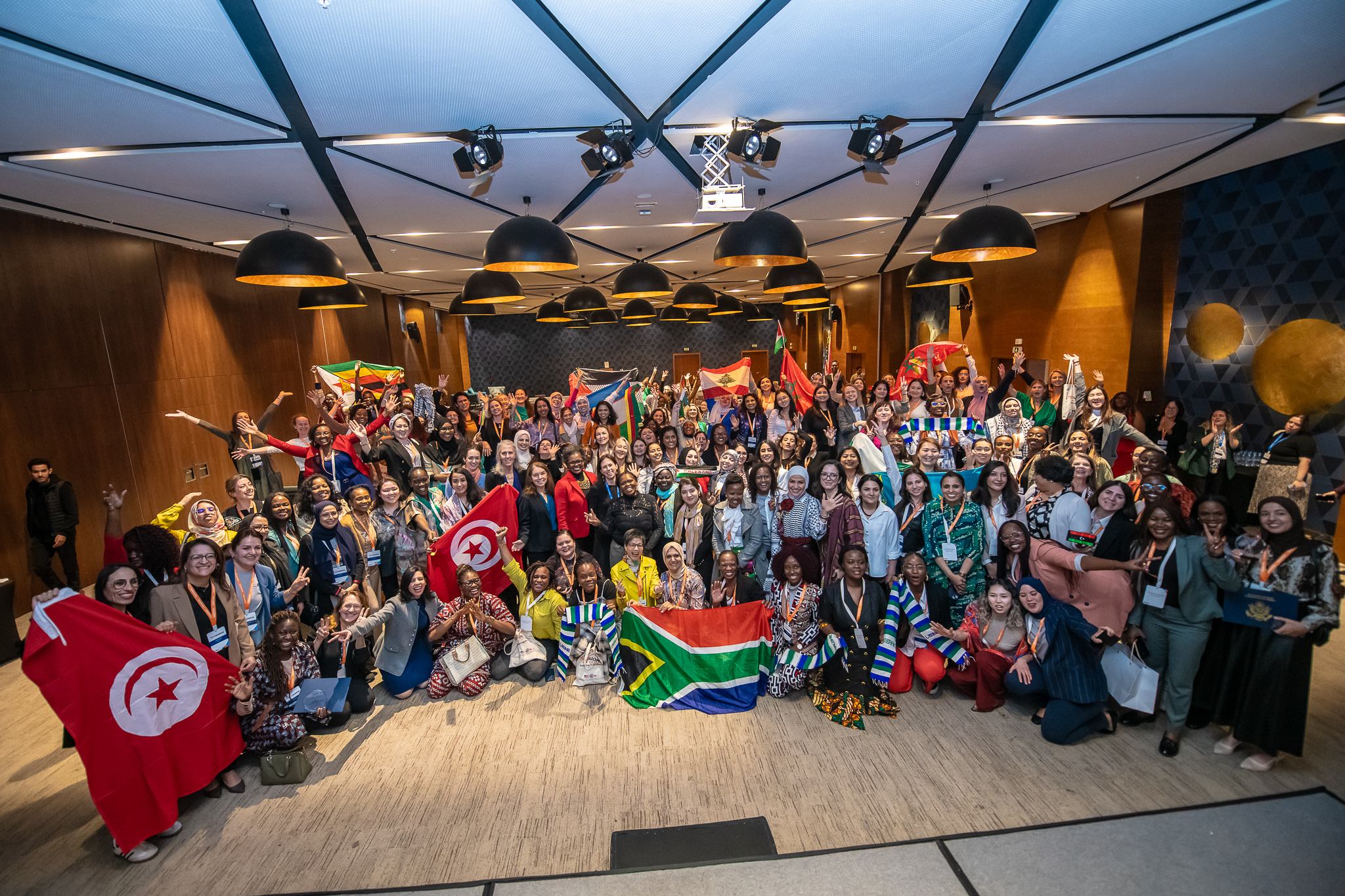 Image of large crowd of people in one room, some holding flags of different nations. Image shows the closing ceremony of the inaugural TechWomen alumn ae summit.