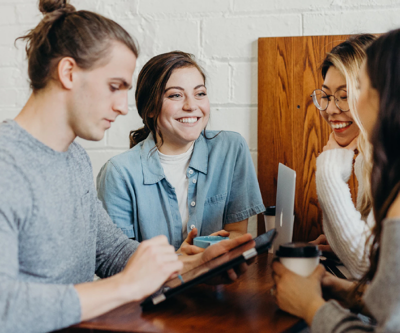 Four people sitting around a table, smiling and engaging in conversation, with a laptop and tablet present.